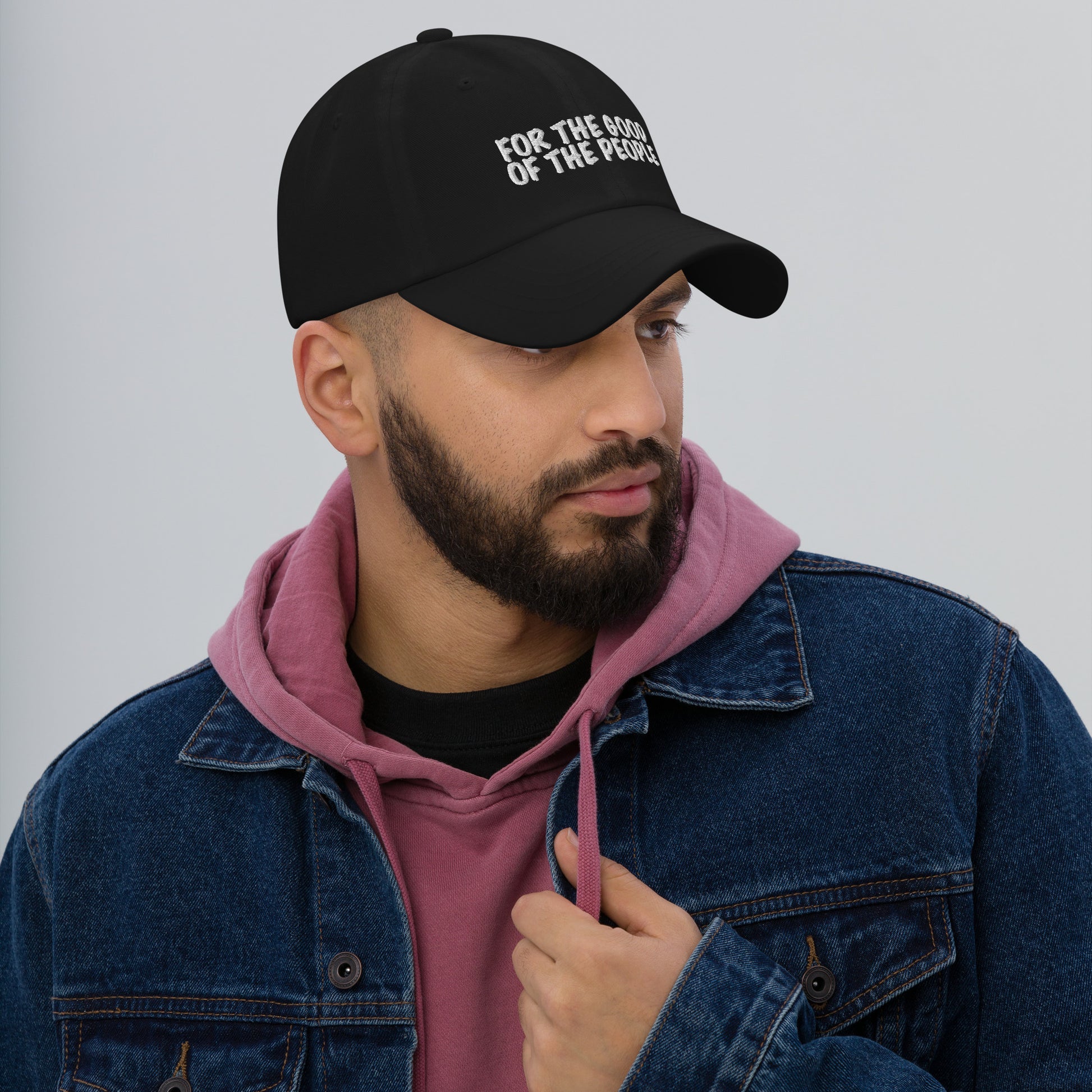 Man wearing blue jean jacket over red sweatshirt with black dad hat with for the good of the people embroidered in white on the front of hat.